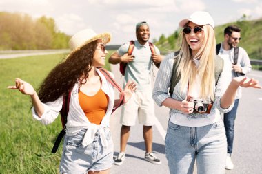 Çok uluslu bir grup arkadaş otobanda yürüyor, birbirleriyle sohbet ediyor, gülüyor, eğleniyorlar. Otostop, yaz macerası, otostop yolculuğu, turizm konsepti.