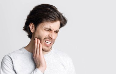 Tooth disease. Man feels pain, grimaces and press hand to cheek isolated on gray background, studio shot clipart