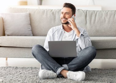 Mesafeli iş ve mobil iletişim. Evden uzakta çalışırken cep telefonuyla konuşan ve dizüstü bilgisayar kullanan, oturma odasındaki kanepenin yanında oturan mutlu serbest çalışan adam.