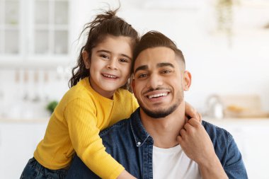 Orta Doğulu mutlu bir baba ve küçük bir kızın mutfak içinde poz verirken, sevimli küçük bir kızın genç babasına sarılırken ve kameraya gülümserken, baba ve çocuğun evde eğlenirken portresi.