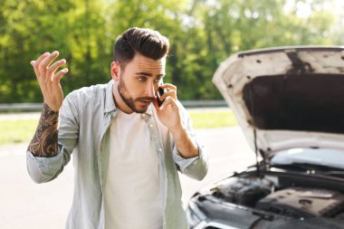 Bozuk kapüşonlu, bozuk arabasının yanında duran, motor arızası yaşayan, cep telefonu konuşmaları yapan, cep telefonuyla bozuk yol servisini arayan genç beyaz bir adam.