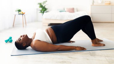 Şişman siyahi kadın karın kası egzersizi yapıyor, köprü yoga pozisyonunda duruyor, evde çekirdek kaslarını çalıştırıyor, uzayı kopyalıyor. Tombul milenyum kadını evde çalışıyor, kilo veriyor.