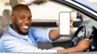 Genç, gülümseyen Afro-Amerikan bir adamın cep telefonunu beyaz ekranla göstererek yeni lüks bir arabanın sürücü koltuğunda oturup, elinde bir aleti tutarak cihaza odaklandığı yakın plan.