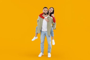 A young couple expresses love and happiness as the woman playfully rides on the mans back. The cheerful yellow background highlights their joyful connection and togetherness. clipart