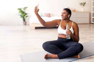 Artı boyut siyahi kadın evde eğitim molasında selfie çekiyor, evde kişisel koçla görüntülü sohbet ediyor. Akıllı telefonlu şişman Afro kadın idmandan sonra kendi fotoğrafını çekiyor.