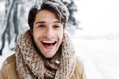 Saçları karlı yakışıklı adam kışın ormandaki kameraya gülümsüyor..