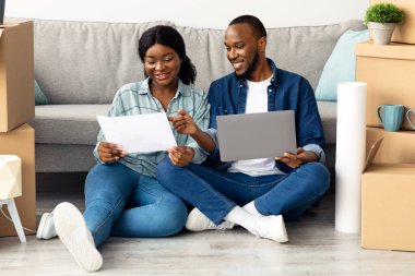 Planları ve dizüstü bilgisayarlı mutlu siyah çift taşındıktan sonra yeni bir daire tasarladı, bilgisayar ve inşaat planı olan Afrikalı genç eşler karton kutuların arasında oturuyor.