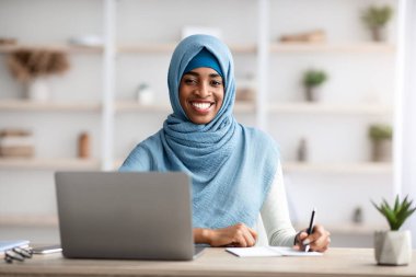Müslüman Kadınlar İçin İş Fırsatları. Bilgisayarla masada oturan, tesettürde gülümseyen, tesettürde çalışan, modern ofiste dizüstü bilgisayarda çalışan ve boş alan alan alan alan arayan mutlu siyah, İslami kadın.