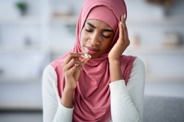Başı ağrıyan ya da evde migren olan hasta siyahi Müslüman kadın, şakakları ovuşturan ve akut ağrıyı azaltmak için ağrı kesici alan Afrikalı kadını üzüyor, boş alanla yakın çekim yapıyor.