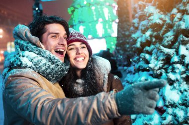 Noel arifesi. Mutlu adam parmağıyla sevgilisini işaret ediyor. Kışın şehir dışında gezen bir şeyi gösteriyor. Geceleri ışıklarla süslenmiş.