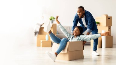 Neşeli Afrikalı Amerikalı çift, yeni dairelerinde eğleniyor, neşeli siyah kadın, parlak oturma odasında karton kutuyla geziyor ve kendi dairelerine taşınmalarını kutluyorlar.