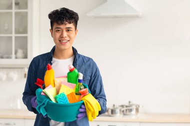 Gülümseyen Asyalı adam temizlikçi, sprey sprey şişesi ve diğer temizlik aletleri, mutfak içi beyaz, fotokopi odası. Neşeli genç Çinli adam hafta sonu ev temizliği yapıyor.