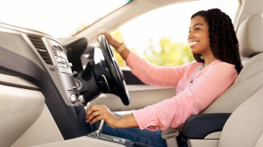 Modern araçta oturan genç Afrikalı Amerikalı kadının profil yan görüntüsü, yeni otomobil kullanan, direksiyon ve vites kolu tutan deneyimli kadın sürücü, yola bakıyor, seçici bir şekilde ele odaklanmış.