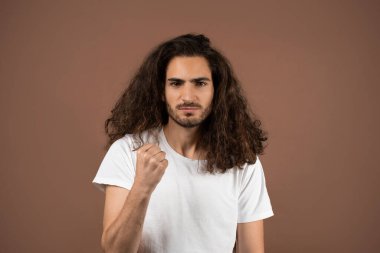 Kıvırcık, uzun saçlı genç bir adam kahverengi bir arka plana karşı güvenle ayakta duruyor. Güçlü bir ifade sergileyerek, azim ve gücü somutlaştıran bir yumruk kaldırır..