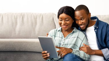 Siyahi Mutlu Milenyum Çifti, evde dinlenirken dijital tablet kullanıyor, gülümseyen Afrikalı Amerikalı eşler online alışveriş yapıyor ya da oturma odasında dinlenirken sosyal ağlara göz atıyorlar.