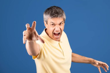 50 'li yaşlarda orta yaşlı bir adam yoğun duygular ifade ediyor. Mavi arka planı olan, sarı tişörtlü ve elini ileri uzatmış bir stüdyoda görünüyor..