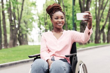 Tekerlekli sandalyedeki mutlu zenci kadın parkta cep telefonuyla selfie çekiyor. Binlerce yıllık Afro-Amerikan kadın engelli, dışarıda yürürken fotoğrafını çekiyor.