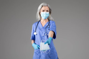 A middle aged woman in scrubs is presenting safety masks with gloved hands. Her calm demeanor underscores the importance of health precautions in a medical environment during crucial times. clipart