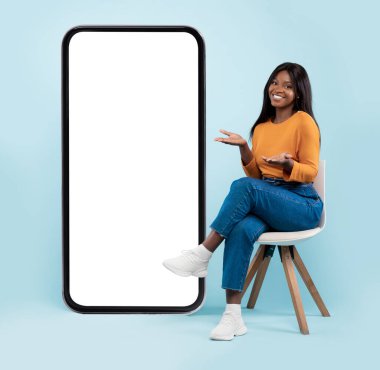 Mobil uygulama ya da web sitesi tasarımı için tasarlanmış büyük boş akıllı telefonun yanında oturan güzel siyahi bir kadın. Mavi stüdyo arka planı, online reklamınız için kopya alanı gösteriyor.