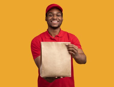 Hızlı Teslimat. Kırmızı şapkalı ve üniformalı Afro-Amerikan kuryenin restorandan kameraya, sarı turuncu stüdyo arka planında el işi kağıt torba taşıdığı ve verdiği ön manzara.