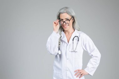 Laboratuvar önlüğü ve gözlük takan orta yaşlı bir kadın doktor güvenle gülümsüyor. Elleri kalçasında duruyor, nötr tonlu bir ortamda pozitif bir davranış sergiliyor, boşluğu kopyalıyor.