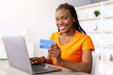 E-ticaret konsepti. Milenyum Afrikalı Amerikalı kadın uzaktan ödeme için kredi kartı kullanıyor, evde laptop üzerinden alışveriş yapıyor. Genç siyahi kadın web mağazasında, kapalı mekanlarda mal satın alıyor.
