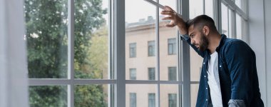 Hayal kırıklığı, kötü hissetmek, kendini izole etmek ve evde yalnız kalmak, derin depresyon, endişe ve agorafobi. Üzgün, genç, sakallı bir adam oturma odasının iç tarafındaki geniş pencereye bakıyor.