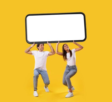 Milenyum çifti, sarı turuncu stüdyo arka planında beyaz ekranlı büyük bir akıllı telefon taşıyor. Mobil uygulama ya da yeni web sitesi reklamları için modelleme, harika bir teklif. Cep telefonu görüntüleme şablonu