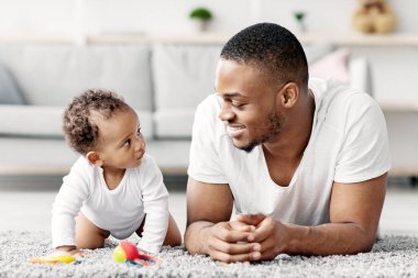 Genç siyah baba ve sevimli bebeği evde birlikte rahatlarken, Afrikalı Amerikalı baba ve tatlı yeni doğan oğlu oturma odasında halıya uzanmış, bebek çocuk babaya merakla bakıyor.