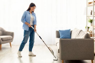 Tam vücut boy görüntüsü, günlük kıyafetlerle gülümseyen genç bir kadın ısıtılmış ahşap kaplama sıcak zemini mikrofiber ıslak bez kullanarak yıkıyor, ev ödevi temizliği yapıyor. Temizlik İşi Konsepti