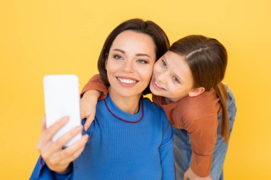 Bir anne ve kızı, parlak sarı bir arka planda selfie çekerken sevgi dolu poz veriyorlar. Kız annesine sarılıyor, ikisi de içten gülümsüyor, aralarındaki bağı ve mutluluğu yakalıyor..