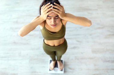 Zayıflatma ve diyet konsepti başarısız oldu. Kilosundan memnun olmayan genç Hintli kadın, terazinin üzerinde duruyor, hayal kırıklığı içinde başını içeride tutuyor, manzaranın üstünde.