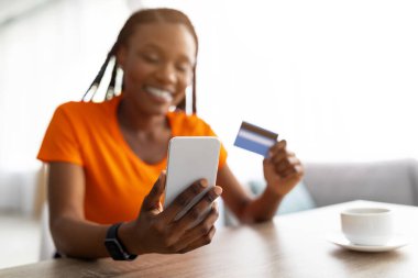 Alışveriş için mobil uygulama. Akıllı telefon ve kredi kartı kullanan neşeli Afro-Amerikan bayan, kafede masada oturuyor, web mağazasından mal alıyor, indirimli ürünler alıyor, seçici odaklanıyor. Boşluğu kopyala