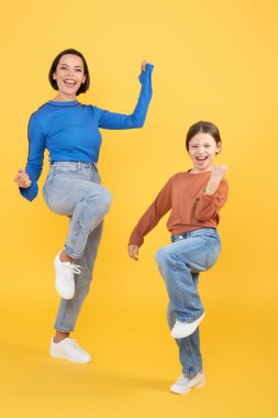 Bir anne ve kızı parlak sarı bir arka planda neşeyle dans ediyorlar. Mutluluk ve sevgiyi ifade ederler. Güçlü bağlarını eğlenceli ve eğlenceli bir hareketle gösterirler..