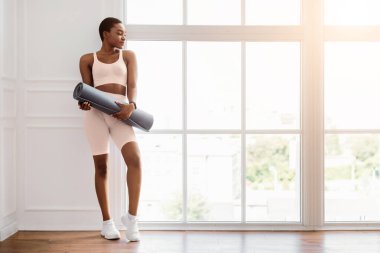 Antrenman için hazır. Afro-Amerikan formda bir kadının spor salonuna giderken portresi, spor salonu veya oturma odasının penceresinde duran paspasları tutarken ve taşırken, boş alana, panorama