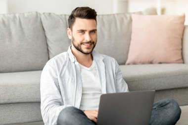 Uzaktan kumandalı iş. Başarılı genç serbest çalışan adam dizüstü bilgisayarda çalışıyor, yere oturuyor ve kanepeye yaslanıyor, evden uzakta çalışıyor, boş bir alanda çalışıyor.