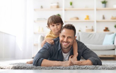 Happy male generations. Indoors portrait of cheerful young dad and his cute little son smiling to camera, lying together on floor at home, free space clipart