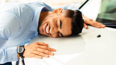 Mutlu genç Arap adam rüyalarının arabasını kucaklıyor, gözlerini kapıyor ve gülümsüyor, yakın plan fotoğraf, yan görüş, kopyalama alanı. Kendinden geçmiş Orta Doğulu bir adam otomobili otomobil salonunu ziyaret ederken bulmuş.