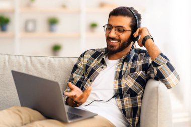 Neşeli, uzun saçlı, sakallı Hintli adam koltukta oturuyor, dizüstü bilgisayar ve kulaklık kullanıyor, evden müşteri ya da kız arkadaşıyla görüntülü görüşme yapıyor, gülümsüyor ve not defterine işaret ediyor, boşluğu kopyalıyor.