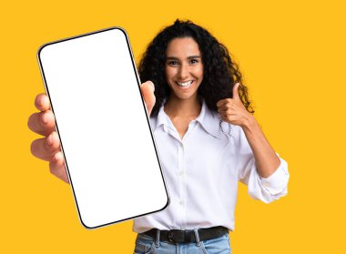 Güzel, orta doğulu, modern cep telefonunu boş ekran ve başparmağıyla gösteren, cep telefonunun boş ekran görüntüsü, modeli, sarı stüdyo arka planında çekilen, boşluğu kopyalayan sevimli bir kadın.