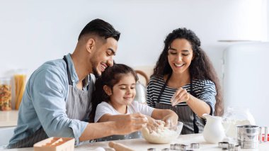 Neşeli bin yıllık Arap ebeveynler küçük kızlarıyla mutfakta kurabiye yapıyorlar cam kasede hamur malzemelerini karıştırıyorlar ve gülüyorlar, baba, anne ve çocuk evde yemek pişirirken eğleniyorlar.