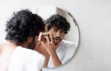 Problem skin. Concerned indian man popping pimple on cheek while standing near mirror in bathroom. Unhappy guy inspecting face, suffering acne, focus on reflection