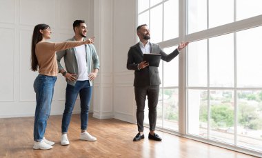 Profesyonel emlakçı, Geniş Daire 'yi Büyük Panoramik Resim Pencereleri' nden güzel manzaraya bakan ve işaret eden genç çifte gösteriyor. Mutlu Adam ve Kadın Ev Sahibi olmaya Hazır