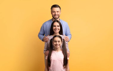 Üç kişilik bir aile, parlak sarı bir arka plana karşı birlikte duruyor. Baba, anne ve kız sevgi ve şefkat gösteriyorlar. Mutlu bir şekilde gülümsüyorlar ve birbirlerine bağlanıyorlar..