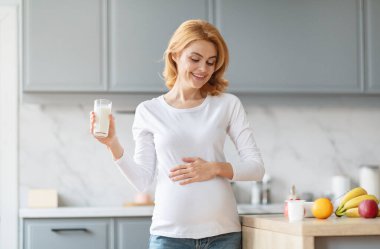 Şık bir mutfakta hamile bir kadın gülümseyerek bir bardak süt tutuyor ve anneliğe olan sevincini gösteriyor. Tezgahta taze meyveler sergileniyor, sağlıklı bir yaşam tarzını ön plana çıkarıyor..