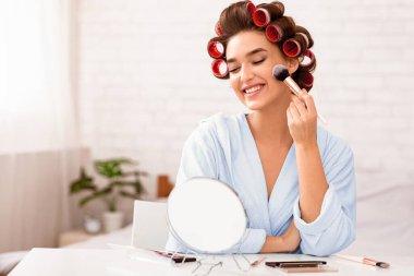 Neşeli kadın yatak odasında makyaj yapıyor, saç bigudileriyle oturuyor ve evde aynada oturan yanaklarına yüz pudrası sürüyor. Kadın Güzelliği Rutini, Günlük Makyaj Konsepti. Boş Alan