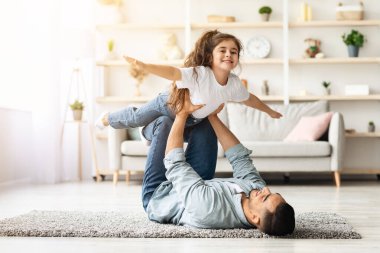 Sevimli küçük kıvırcık kız babasıyla evde vakit geçiriyor. Orta doğulu genç baba tatlı kızıyla uçak oyunu oynuyor. Oturma odasında yere uzanıyor.
