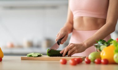 Pembe elbiseli bir kadın ahşap bir tahta üzerinde ustaca salatalık keser. Taze domatesler ve diğer sebzeler onu parlak, çağdaş bir mutfakta, kırpılmış, kopyalayıcı uzayda kuşatıyorlar.