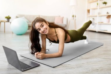 Güçlü Hintli kadın dizüstü bilgisayarın önünde dirsek tahtası üzerinde duruyor, evde spor dersleri veriyor. Karın kaslarını çalıştıran formda bir Asyalı kadın. Covid izolasyonu sırasında ev içi uygunluk
