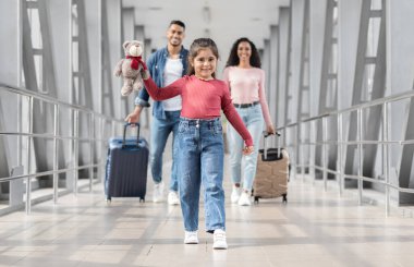 Havalimanında ailesiyle yürüyen neşeli Orta Doğulu bir kızın portresi, birlikte tatile çıkmaya hazır mutlu genç bir Arap aile, Terminalin çıkış kapısına gidiyor, seçici bir şekilde çocuğa odaklanıyor.